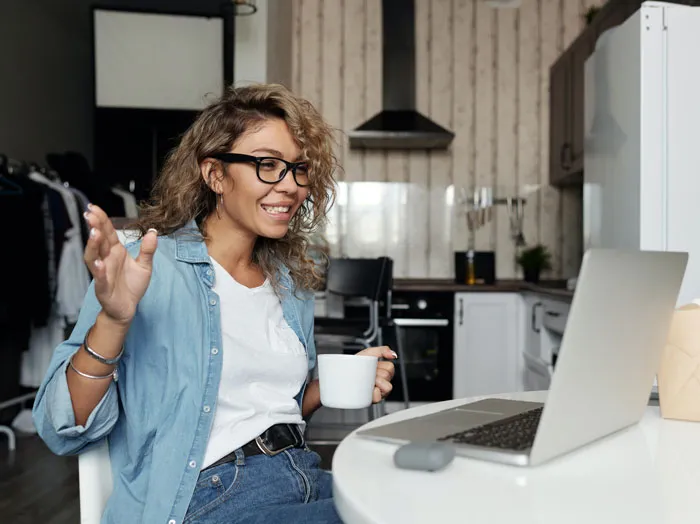 Woman enjoying fun activities at home, smiling with a cup of coffee and engaging in a video call on her laptop.