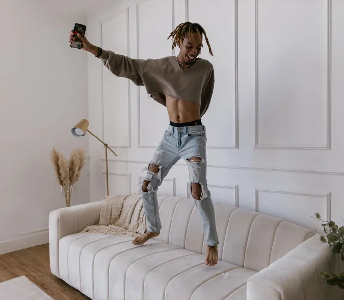 Person joyfully jumping on a couch at home to avoid boredom, holding a phone in hand.