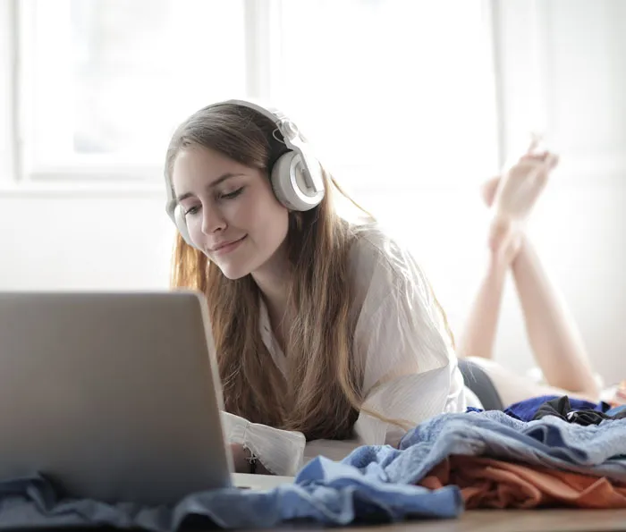 Woman enjoying music with headphones, lying down and using a laptop at home to avoid boredom.