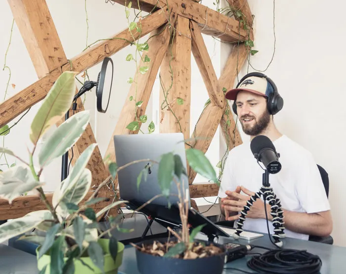 Man enjoying a home podcasting session with a laptop, microphone, and plants, engaging in fun activities to avoid boredom.