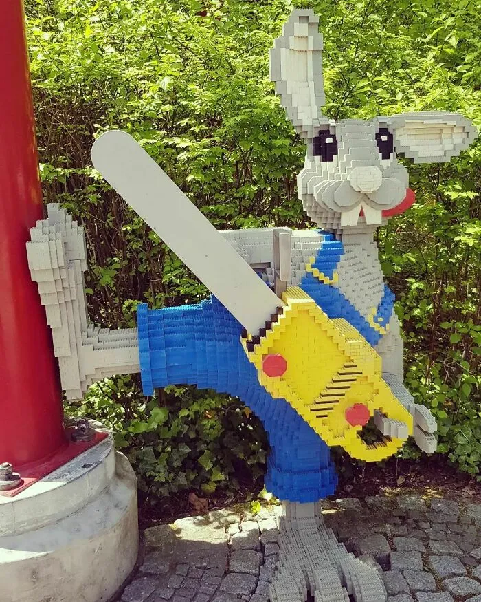 LEGO builds sculpture of a gray rabbit in blue overalls holding a yellow chainsaw, set outdoors near green bushes.