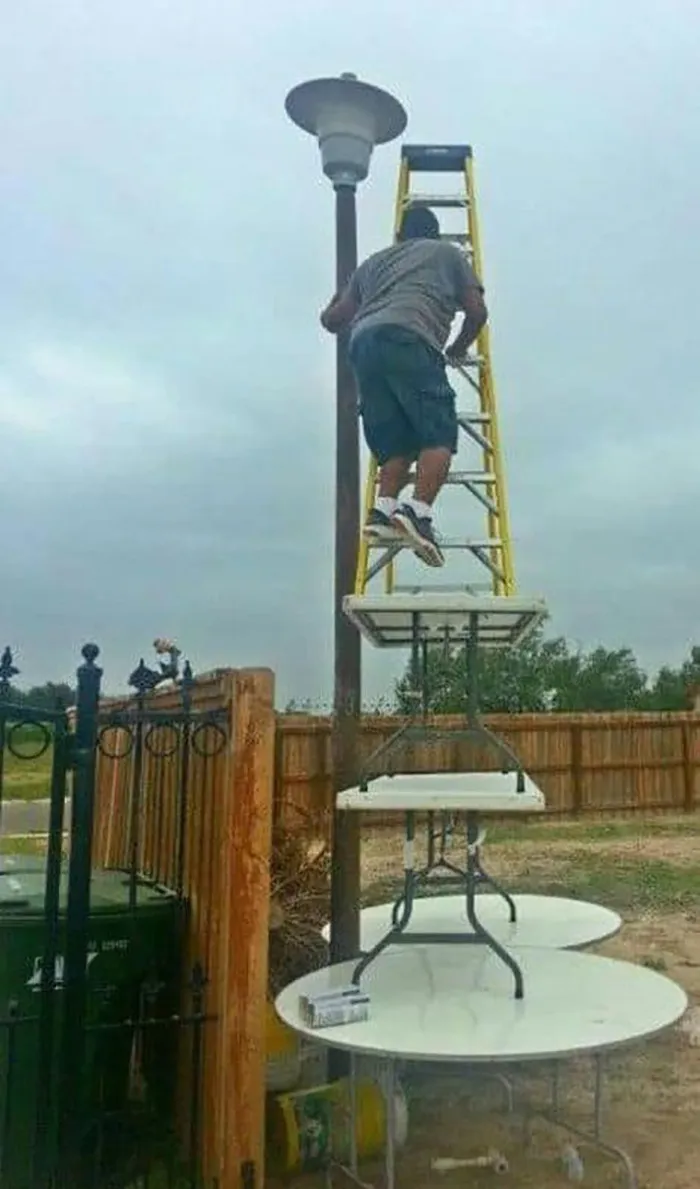 Person on a ladder balancing on stacked tables, defying OSHA safety regulations to fix a streetlight.