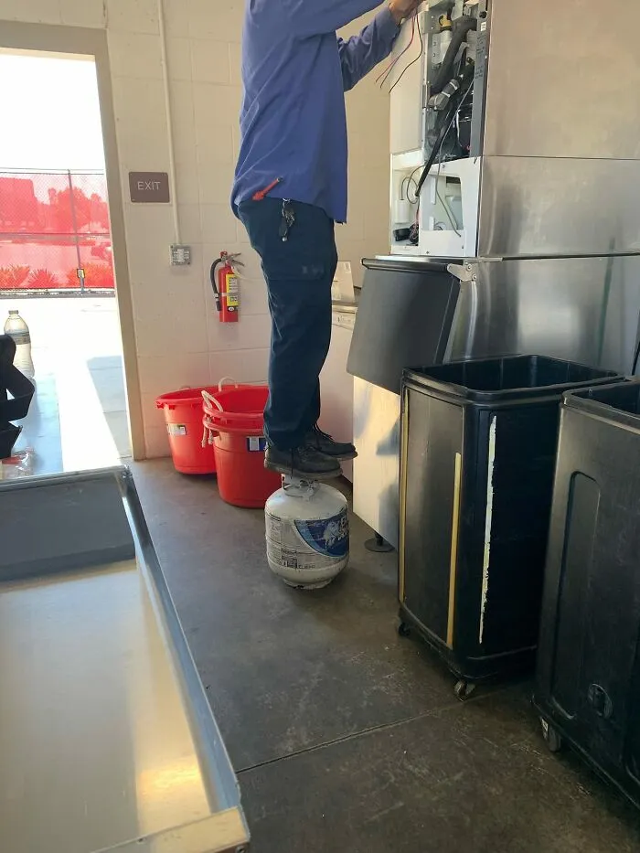 Person standing on a propane tank for balance while fixing machinery, ignoring OSHA safety regulations.