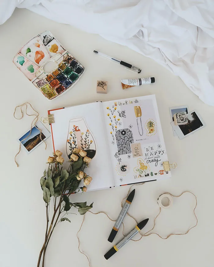Person enjoying me time by painting in a book, surrounded by art supplies and dried flowers.