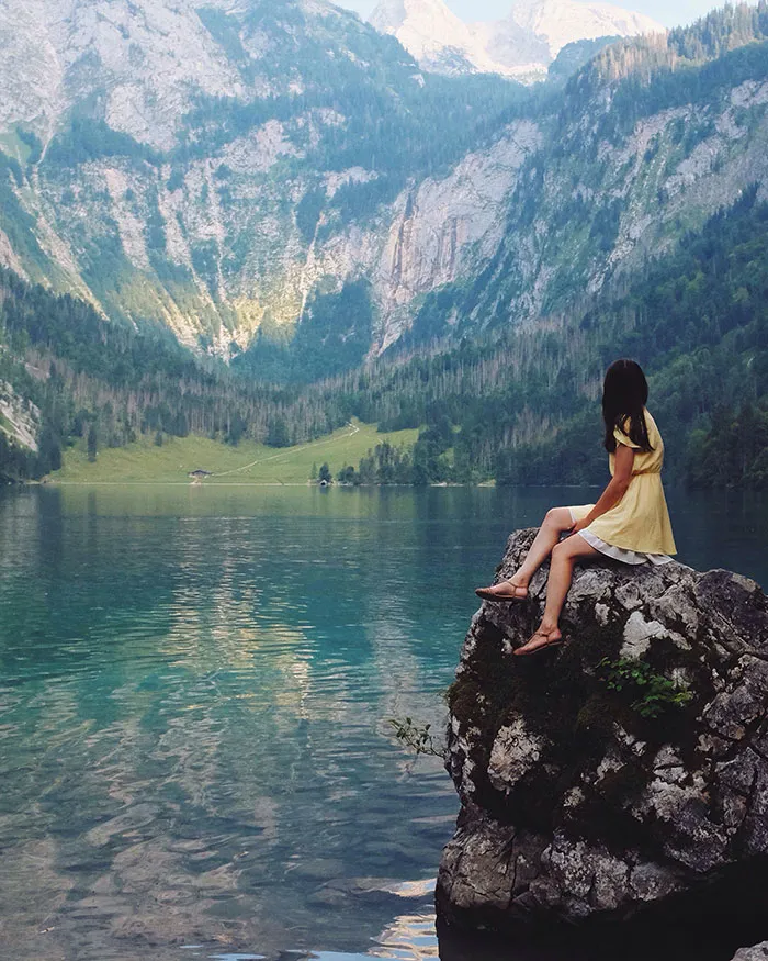 Girl sitting on a rock by a serene lake, gazing at majestic mountains, enjoying some me time.
