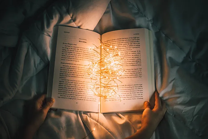 Person enjoying me time, holding a book with glowing fairy lights on a cozy bedspread.