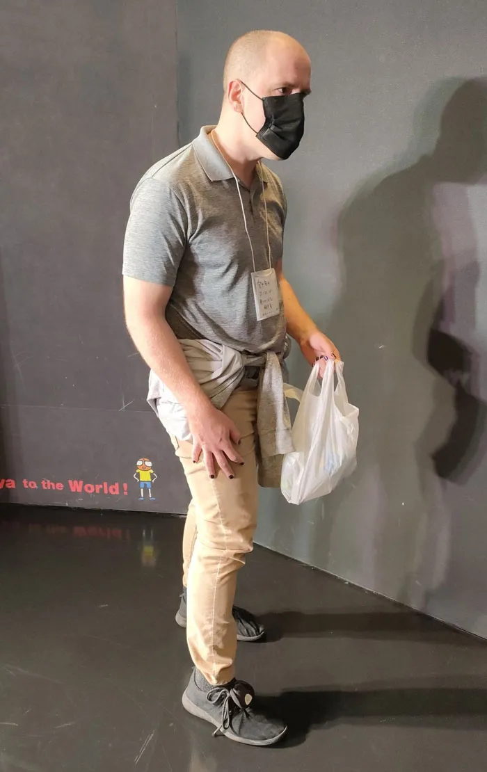 Man in simple costume for Mundane Halloween, wearing a mask and holding a shopping bag, stands against a dark wall.