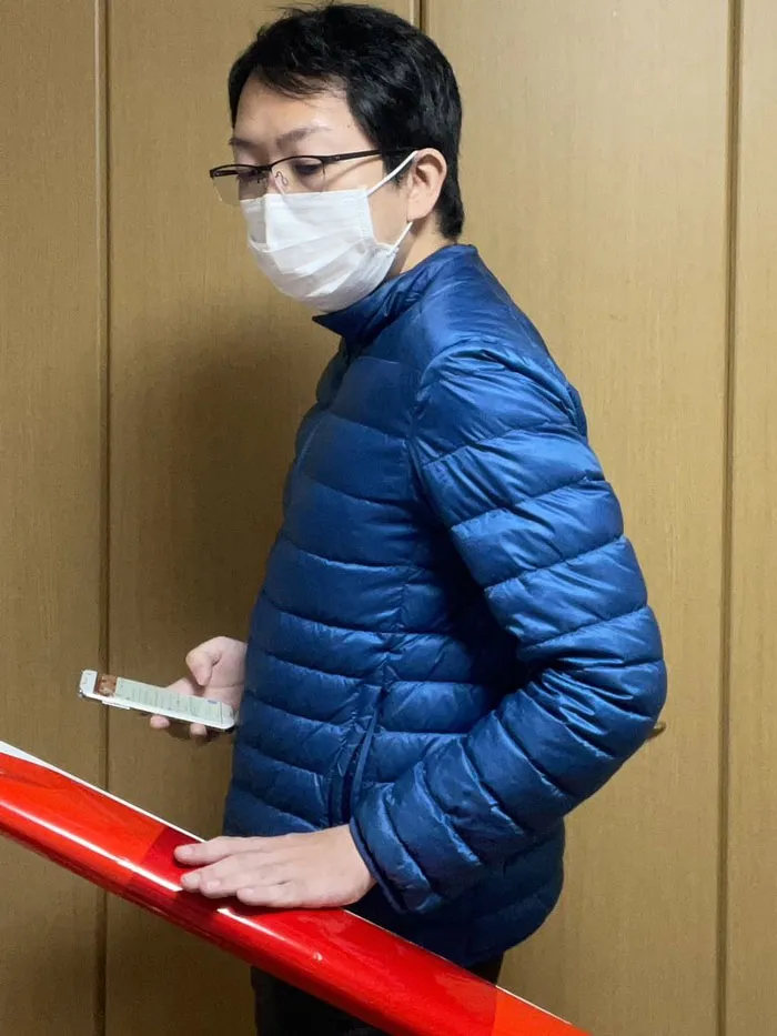 Person in a mundane Halloween costume, wearing a mask and blue jacket, looking at a phone while standing by a handrail.