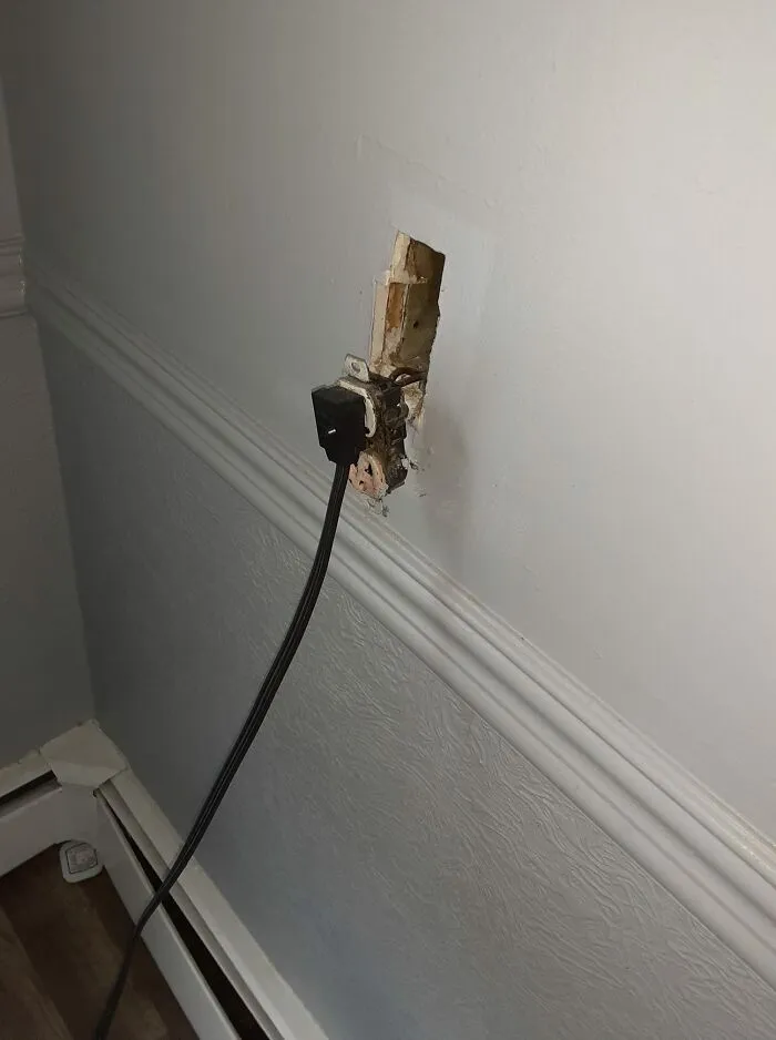 Unsafe electrical socket setup with exposed wires against a wall, ignoring OSHA safety regulations.