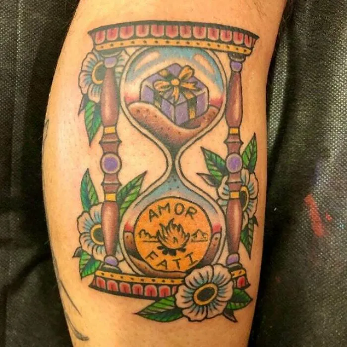 Trippy tattoo of an hourglass with flowers, a gift, and "Amor Fati" text, mixing vibrant colors and intricate design.