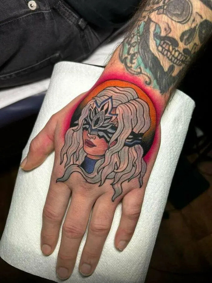 Trippy tattoo of a masked woman with flowing hair on a hand, featuring vibrant colors and abstract design.