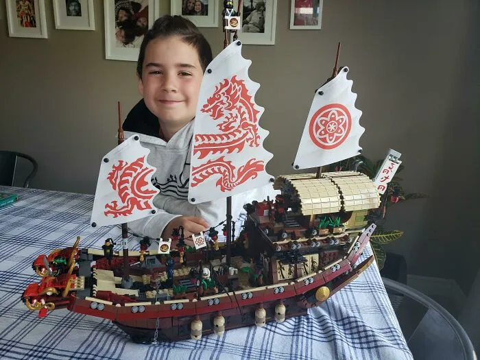 Child smiling behind an intricate LEGO build of a detailed ship with decorated sails, showcasing incredible LEGO builds art.