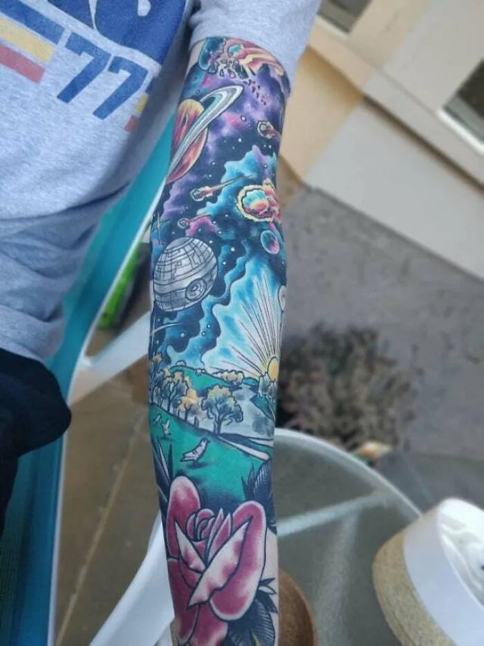 Colorful trippy tattoo of a cosmic scene with planets, stars, and a rose on an arm.
