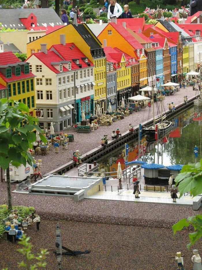 Colorful LEGO builds of detailed city buildings and waterfront scene with miniature figures along a canal reflecting art.