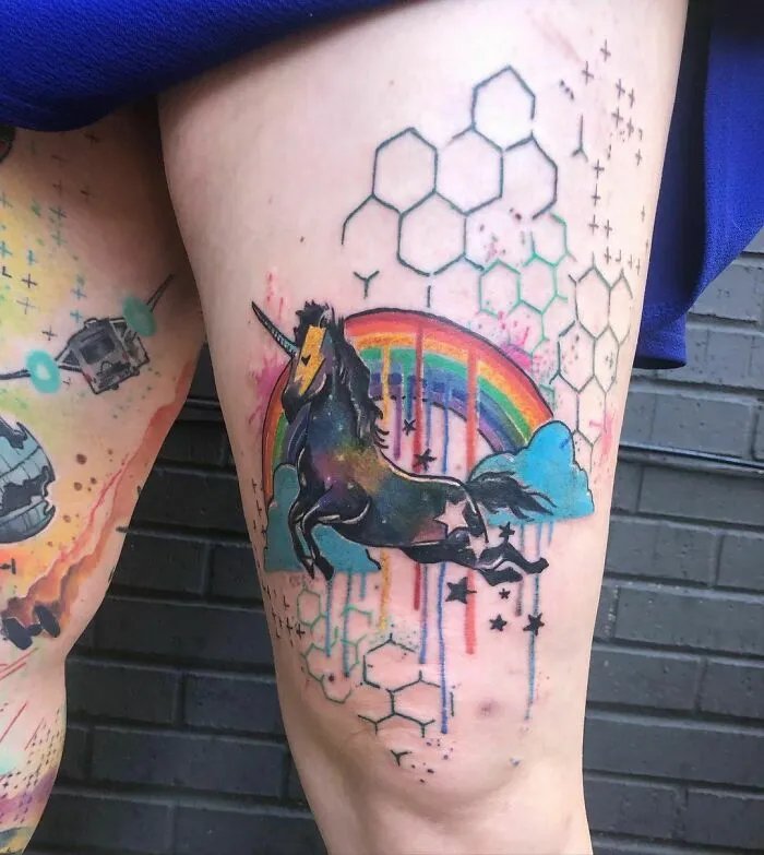 Tattoo of a psychedelic unicorn with a rainbow on a thigh, featuring geometric patterns and vibrant colors.