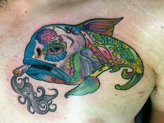 Colorful trippy tattoo of a psychedelic fish with unique lettering and a cigarette on a person's chest.