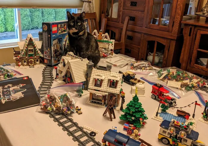Cat sitting among intricate LEGO builds on a table, featuring detailed houses, a Christmas tree, and train tracks.