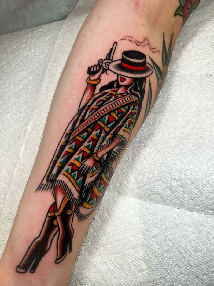 Colorful trippy tattoo of a mysterious figure wearing a patterned poncho and hat, holding a revolver on a person's forearm.