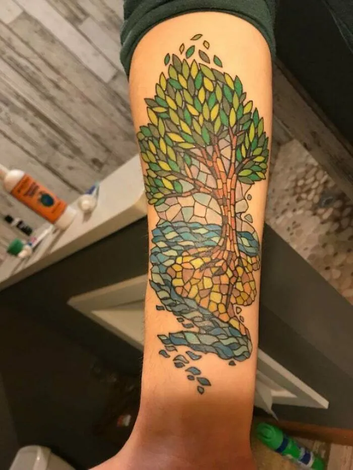 Colorful trippy tattoo of a tree with mosaic style leaves and water on a forearm, creating an optical illusion.