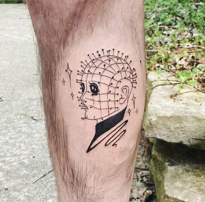 Trippy tattoo of a head with grid lines and stars, on a person's leg, surrounded by greenery and rocks.