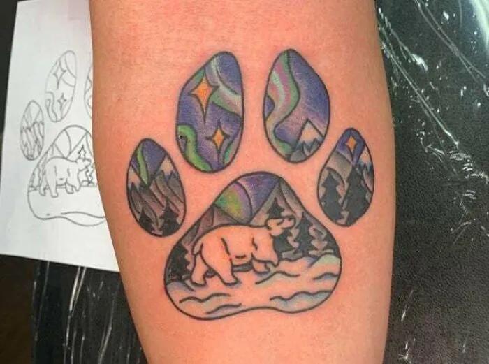 Trippy tattoo of a bear in a paw print design with a colorful aurora and mountain scenery.