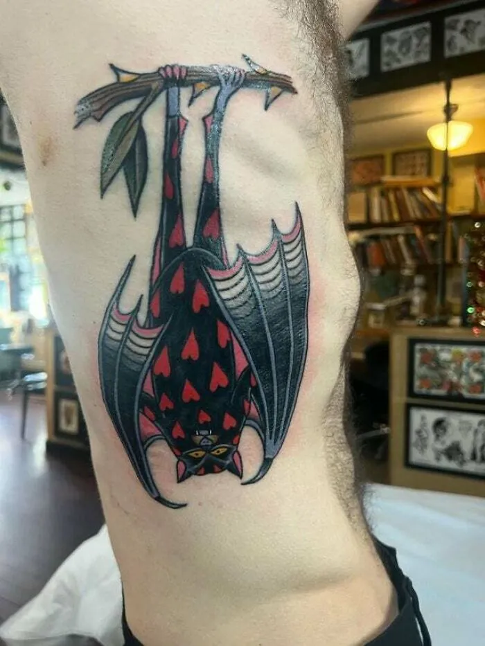 Trippy tattoo of a geometric bat with heart patterns hanging upside down on an arm, surrounded by vibrant studio decor.