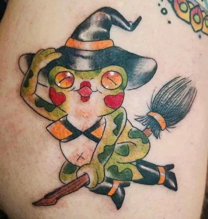 Trippy tattoo of a cartoon frog dressed as a witch, flying on a broomstick with a quirky expression, using bold colors.