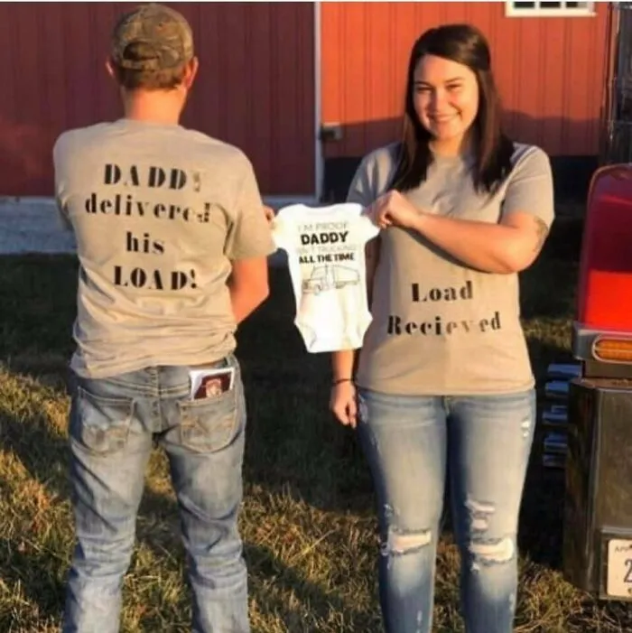 Couple in matching shirts with humorous text, holding a baby onesie, causing cringe-worthy moment in front of a barn.
