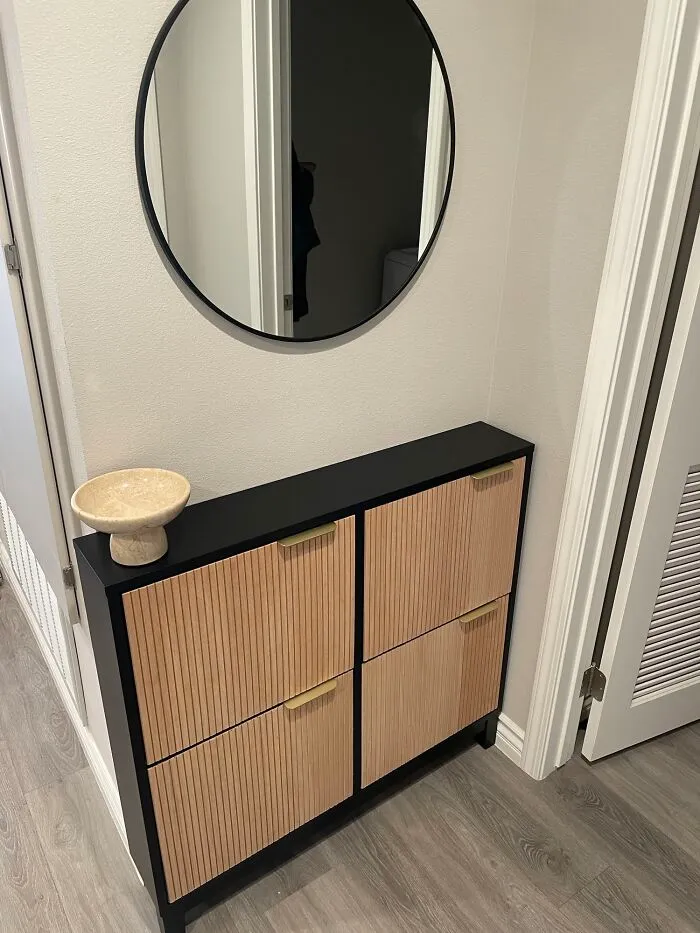 IKEA furniture hack with a minimalist cabinet featuring textured wooden doors and a decorative round mirror above.