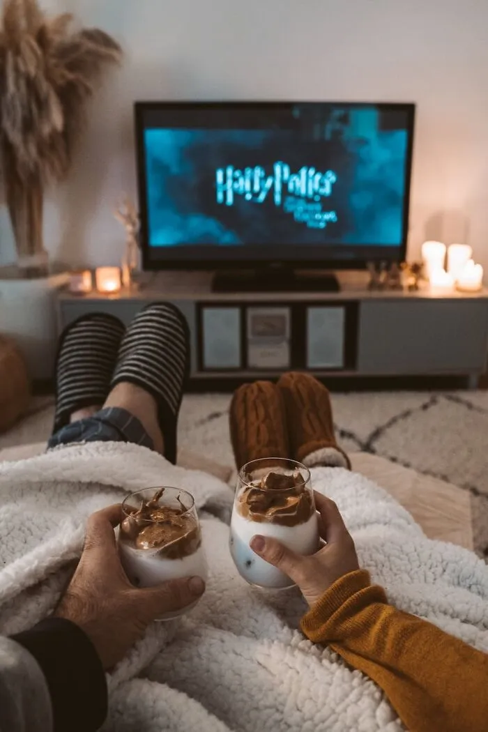 Couple enjoying cozy New Year’s Eve at home with drinks, watching a movie in a warmly lit living room.