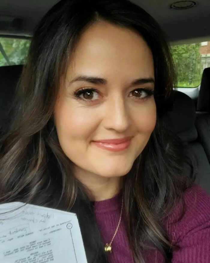 Woman with long dark hair holding a paper, smiling inside a car, representing smartest celebrities and intelligence.