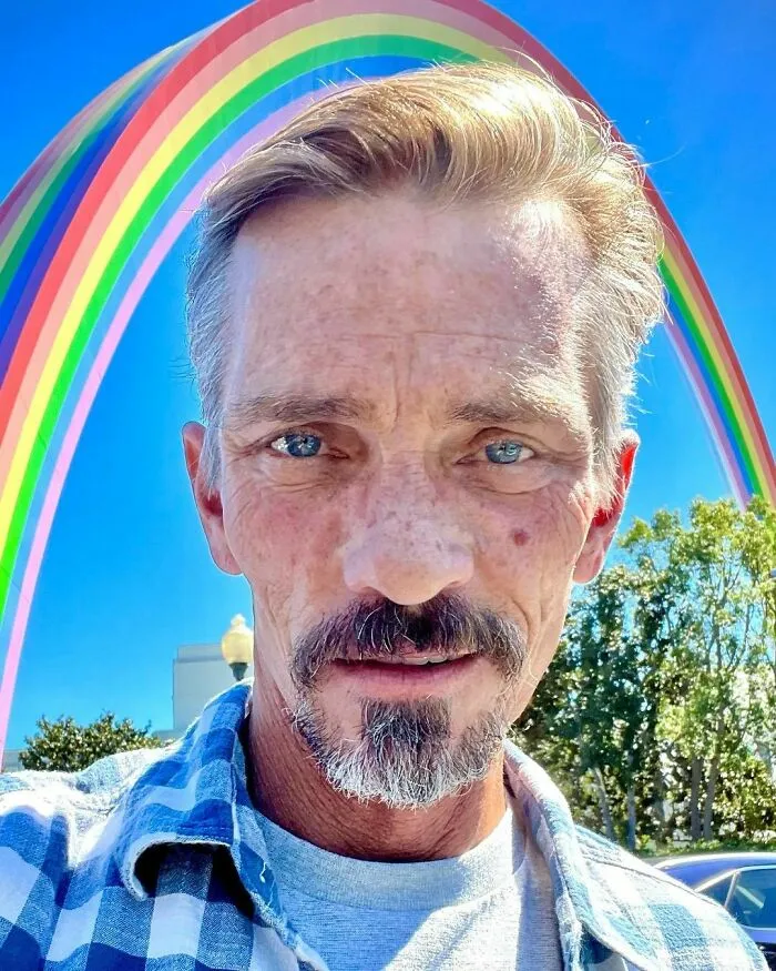 Close-up of a male celebrity with blue eyes and a goatee, standing outdoors under a vibrant rainbow on a sunny day.