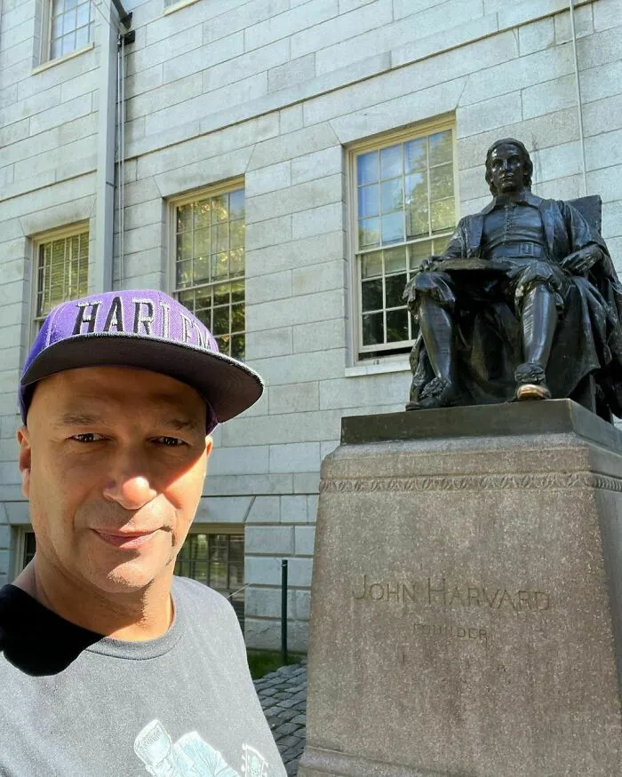 Man wearing a Harlem hat taking a selfie near a historic statue outside a university building, representing intelligent celebrities.