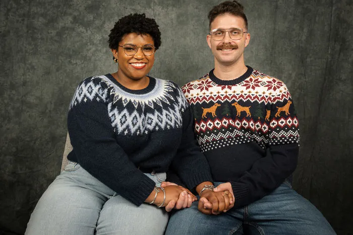 Couple in 80s-themed photoshoot wearing festive sweaters and jeans, sitting and holding hands.