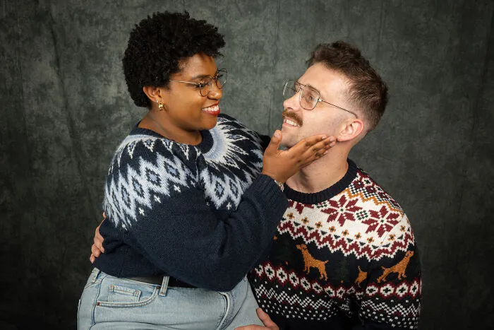 Couple in awkward 80's style photoshoot for anniversary, wearing vintage sweaters with fond expressions.