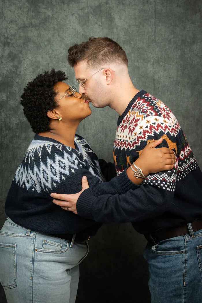 Couple in 80s-themed photoshoot, wearing patterned sweaters and glasses, sharing a kiss for their anniversary.