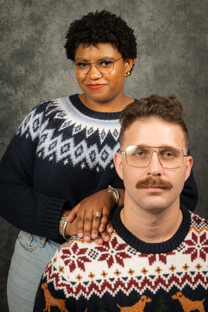 Couple in 80s-themed photoshoot, wearing retro sweaters and glasses, posing for anniversary.