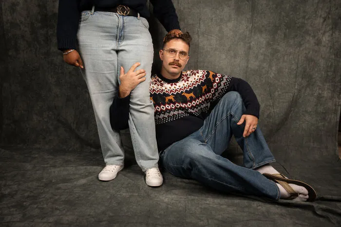 Couple in an awkward 80's themed photoshoot, man seated on the floor, both wearing retro outfits.
