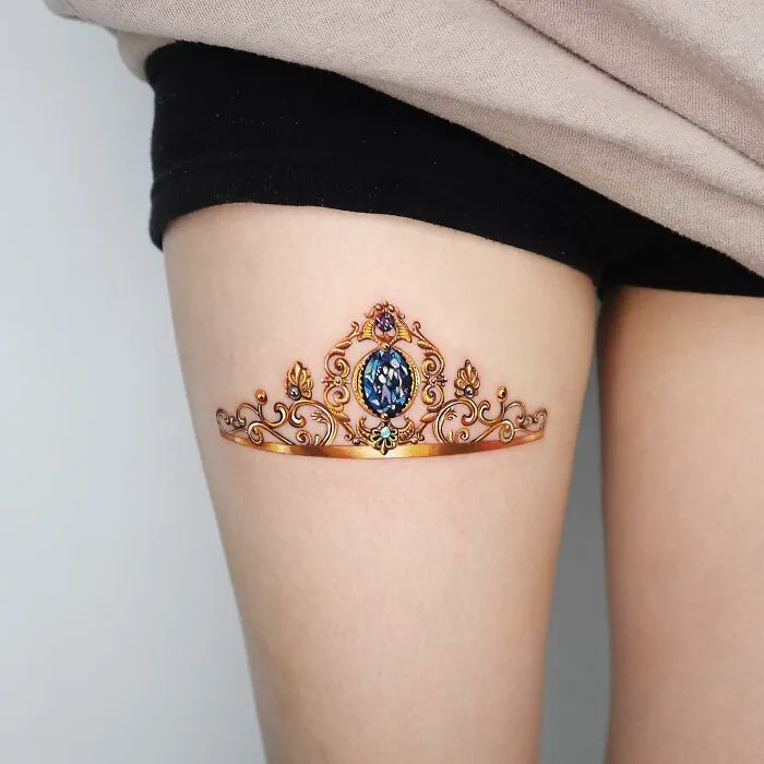 Golden tattoo of an ornate crown on thigh, crafted by Manhattan-based artist.