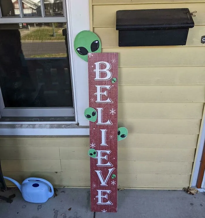 Humorous Christmas decor with aliens peeking around a "Believe" sign on a porch.