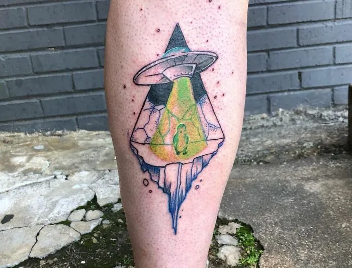 Trippy tattoo of an alien abducting a penguin on a leg, showcasing vibrant colors and surreal design.