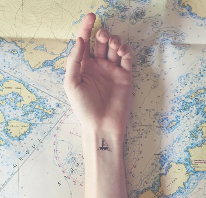 Minimalist tattoo of a sailboat on a wrist, positioned over a nautical map background.
