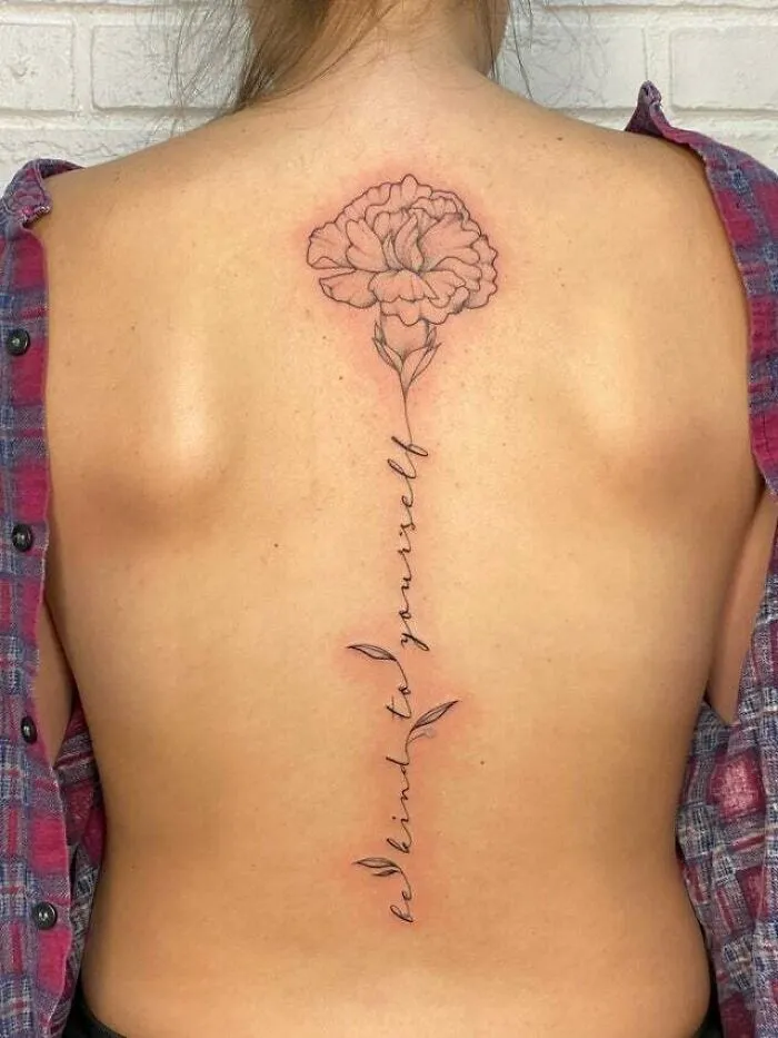 Minimalist tattoo of a flower and "be kind to yourself" text on a person's back, creating an aesthetically pleasing design.