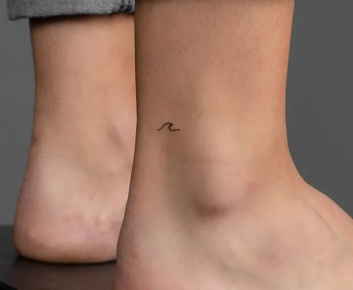 Minimalist ankle tattoo of a small black wave on skin, aesthetically pleasing and simple design.