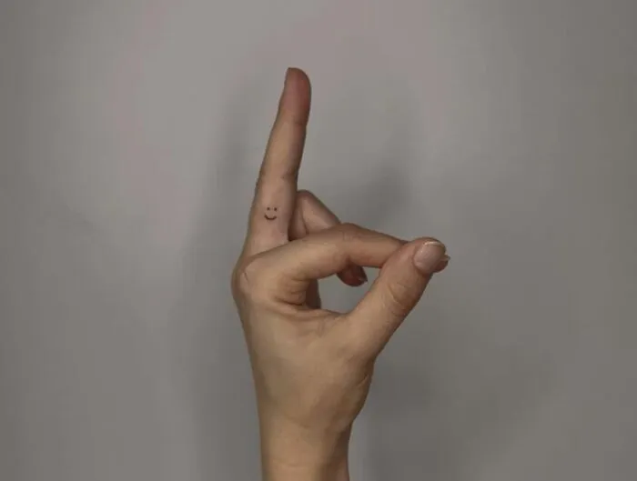 Hand gesture with a minimalist smiley face tattoo on the finger, aesthetically pleasing minimalist tattoo design.