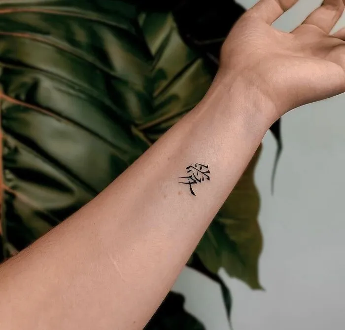 Minimalist tattoo of black Japanese characters on a forearm, stylishly placed against a leafy background.