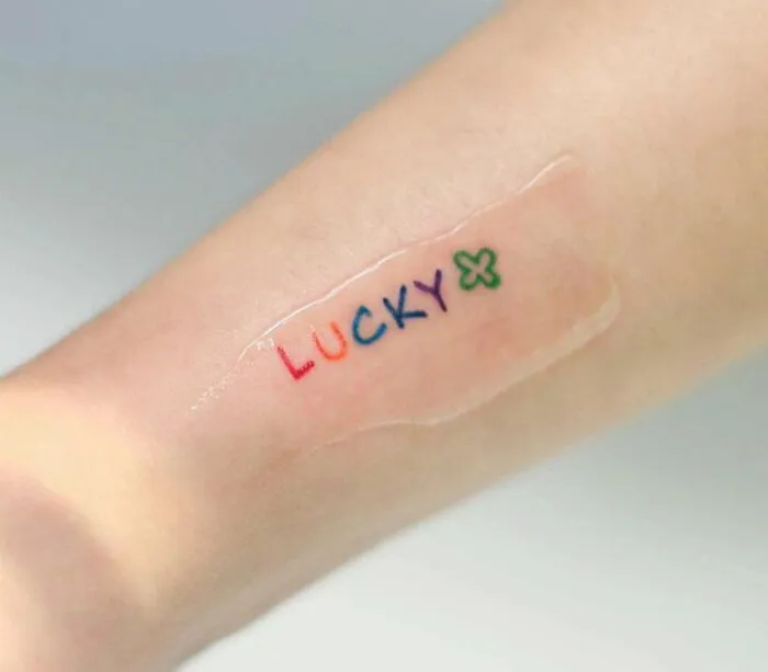 Minimalist tattoo of the word "Lucky" in colorful letters with a green clover design on an arm.