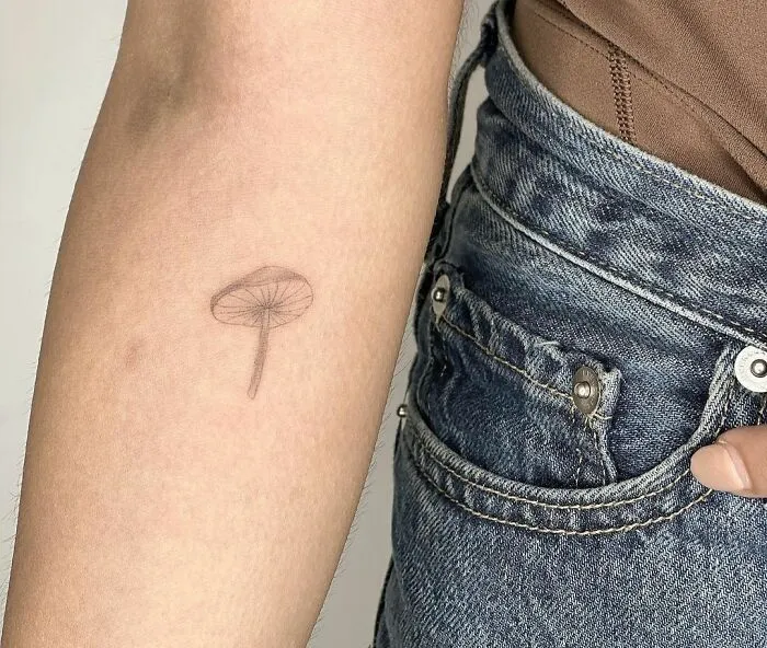 Minimalist tattoo of a small mushroom on a person's forearm, next to blue jeans.
