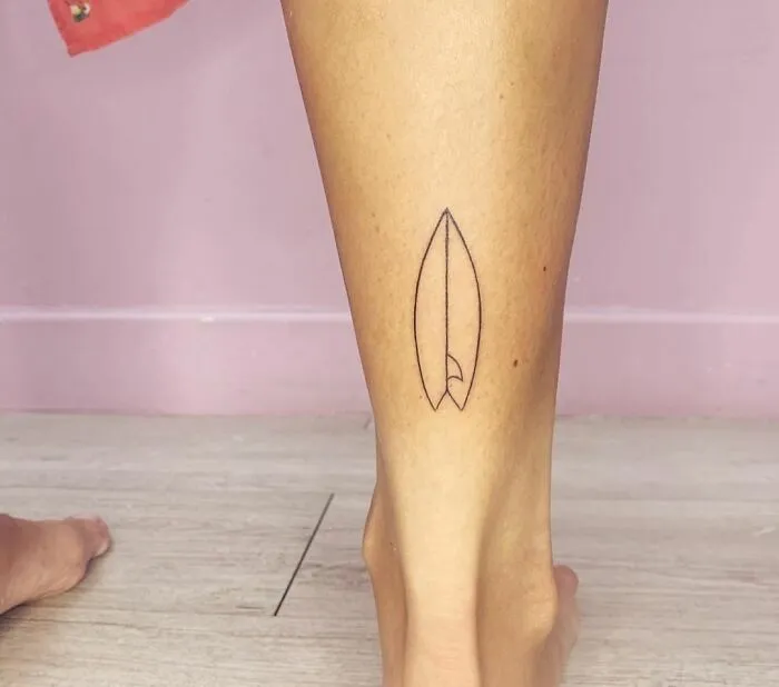 Minimalist tattoo of a surfboard on a person's lower leg, featuring simple black line art.