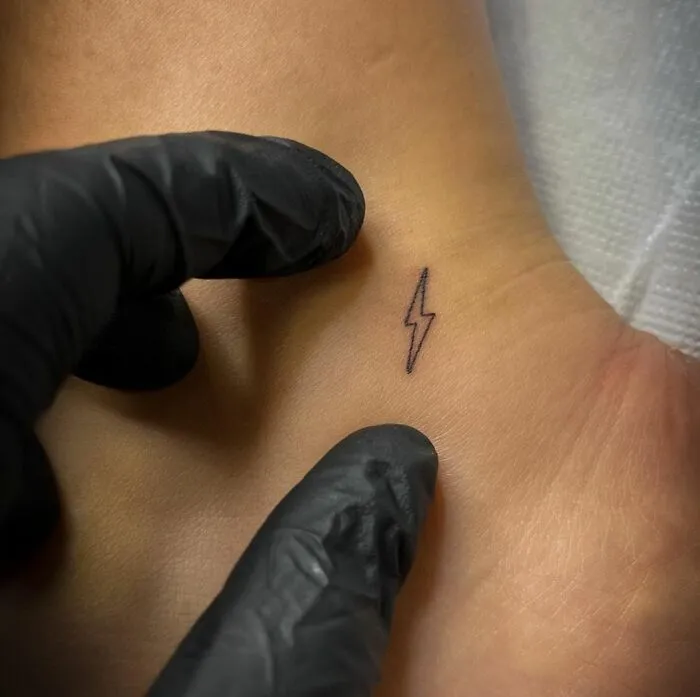Minimalist lightning tattoo on wrist, created with fine lines by gloved hands.
